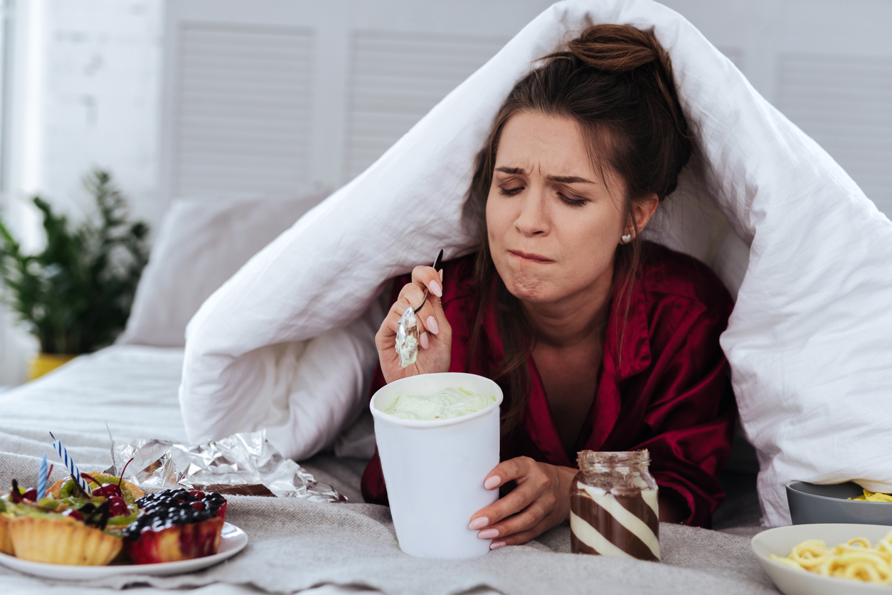 Stressed woman hiding under blanket eating junk food. Learn 3 ways to stop stress eating.