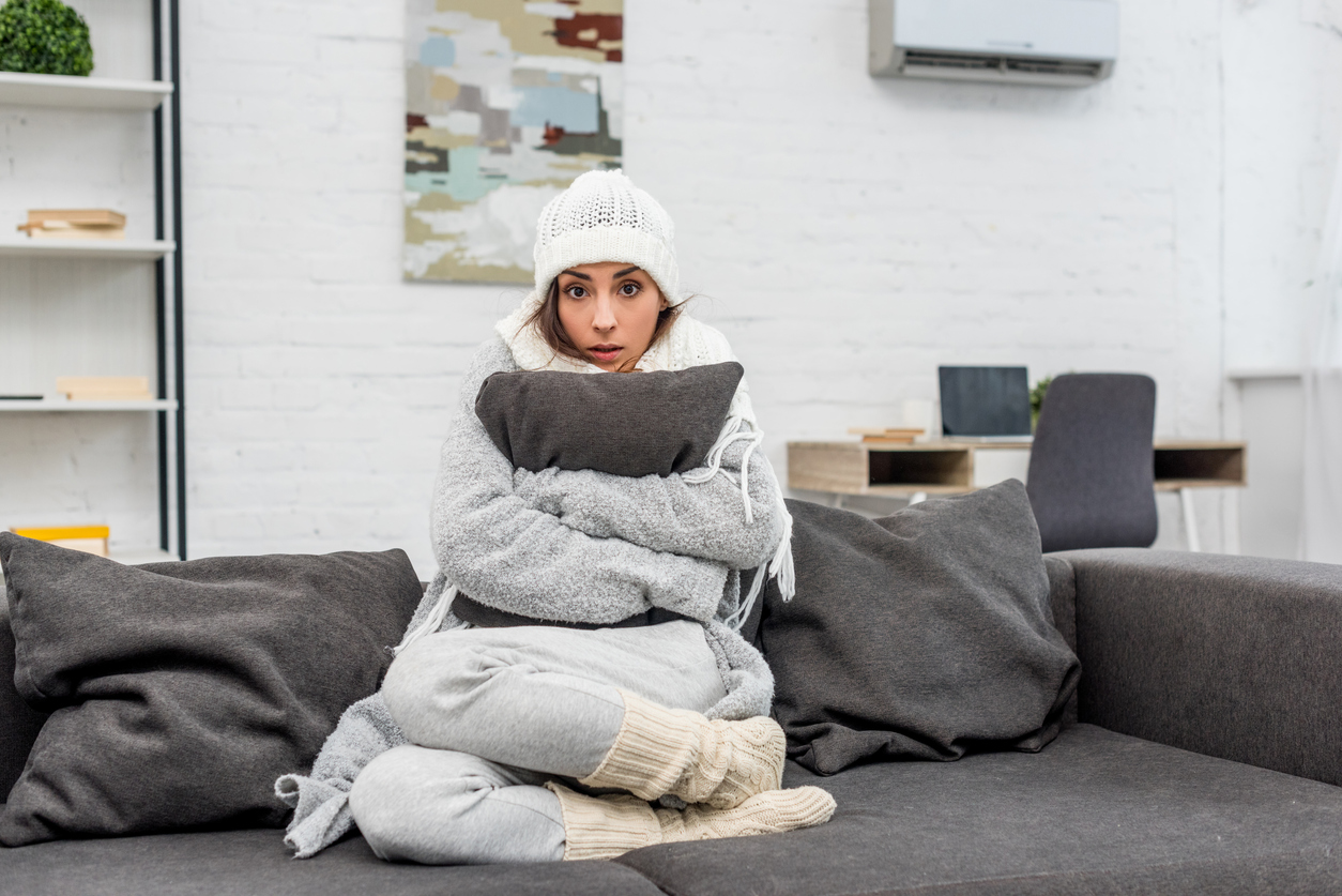 freezin young woman in warm clothes sitting on couch and hugging cushion at home. Learn about cold flashes and menopause.