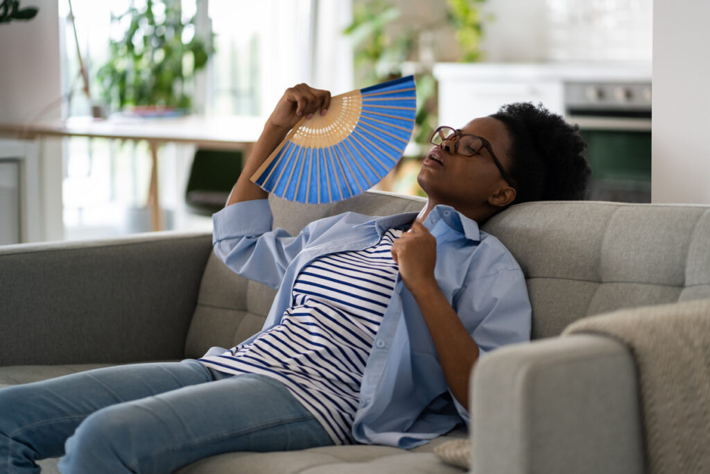 Black woman on couch with fan trying to cool down after menopause night sweat