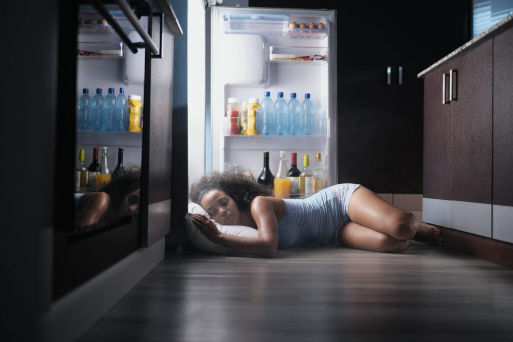 Black woman laying in front of a refridgerator trying to cool down after a night sweat
